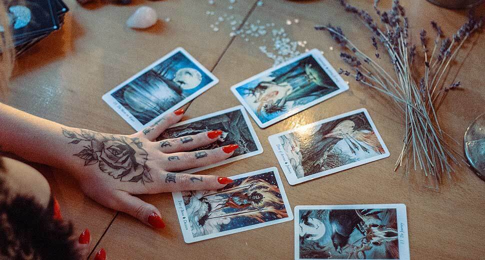 Foto de uma mesa. Sobre ela aparece a mão de uma mulher, tatuada, mexendo em cartas de tarô.